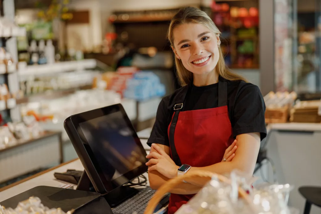 In a modern grocery store environment, a friendly cashier wearing an apron smiles warmly at the customers