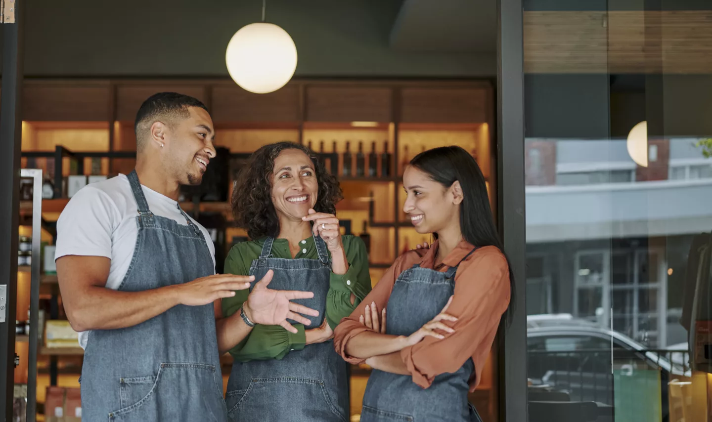 Mature female artisanal deli owner and two young employees smiling and talking together while standing at the entance to her shop