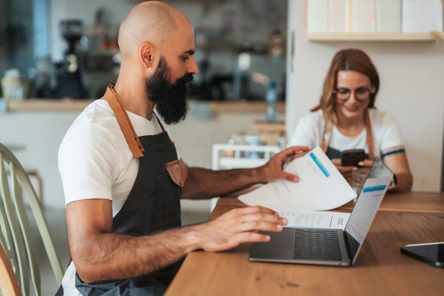Cafe owners in aprons work with a laptop and paperwork, representing small business logistics, order management, finance and teamwork
